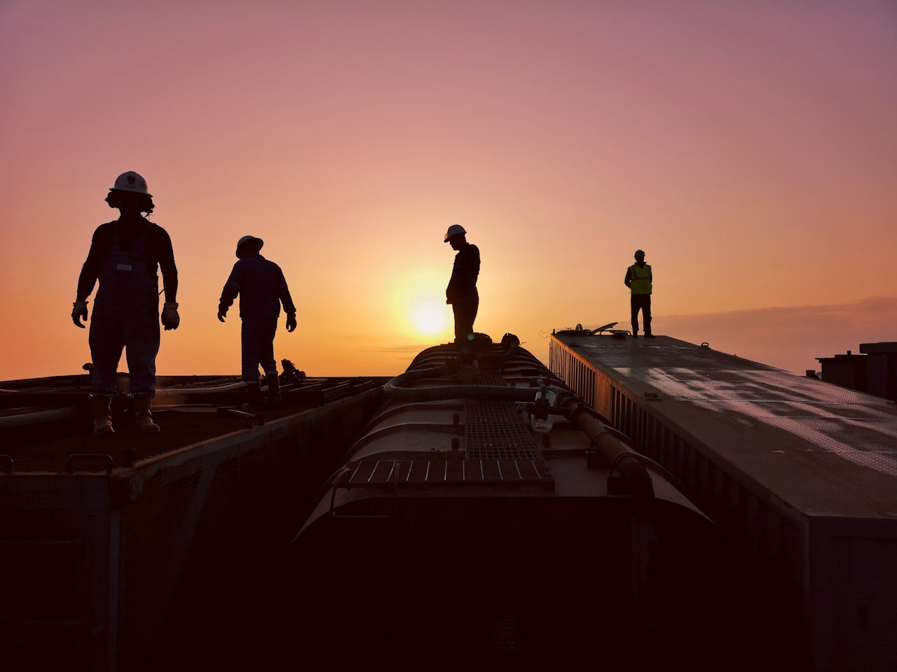 Silhouettes of workers atop industrial structures against a stunning sunset backdrop.